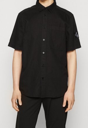 Osoba ubrana w czarną koszulę na krótki rękaw z guziczkami, kieszenią na piersi i naszywką z logo na lewym rękawie, w połączeniu z czarnymi spodniami.