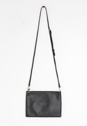 Sac bandoulière rectangulaire en cuir noir avec une fine sangle ajustable et une fermeture éclair sur le dessus, suspendu sur un fond blanc.
