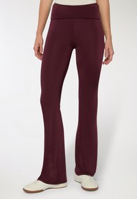 Pantaloni sportivi bordeaux con vita alta, gambe svasate, texture liscia e abbinati a scarpe da ginnastica bianche su uno sfondo neutro.