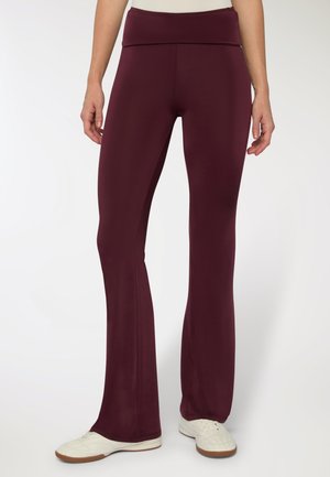 Pantaloni sportivi bordeaux con vita alta, gambe svasate, texture liscia e abbinati a scarpe da ginnastica bianche su uno sfondo neutro.