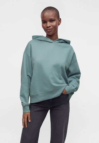 Personne portant un sweat à capuche ample bleu canard et un jean noir, debout avec une main dans la poche devant un fond clair et uni.