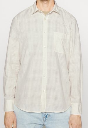Homme portant une chemise beige clair à manches longues avec un motif à carreaux subtil et une poche poitrine, associée à un jean bleu clair.