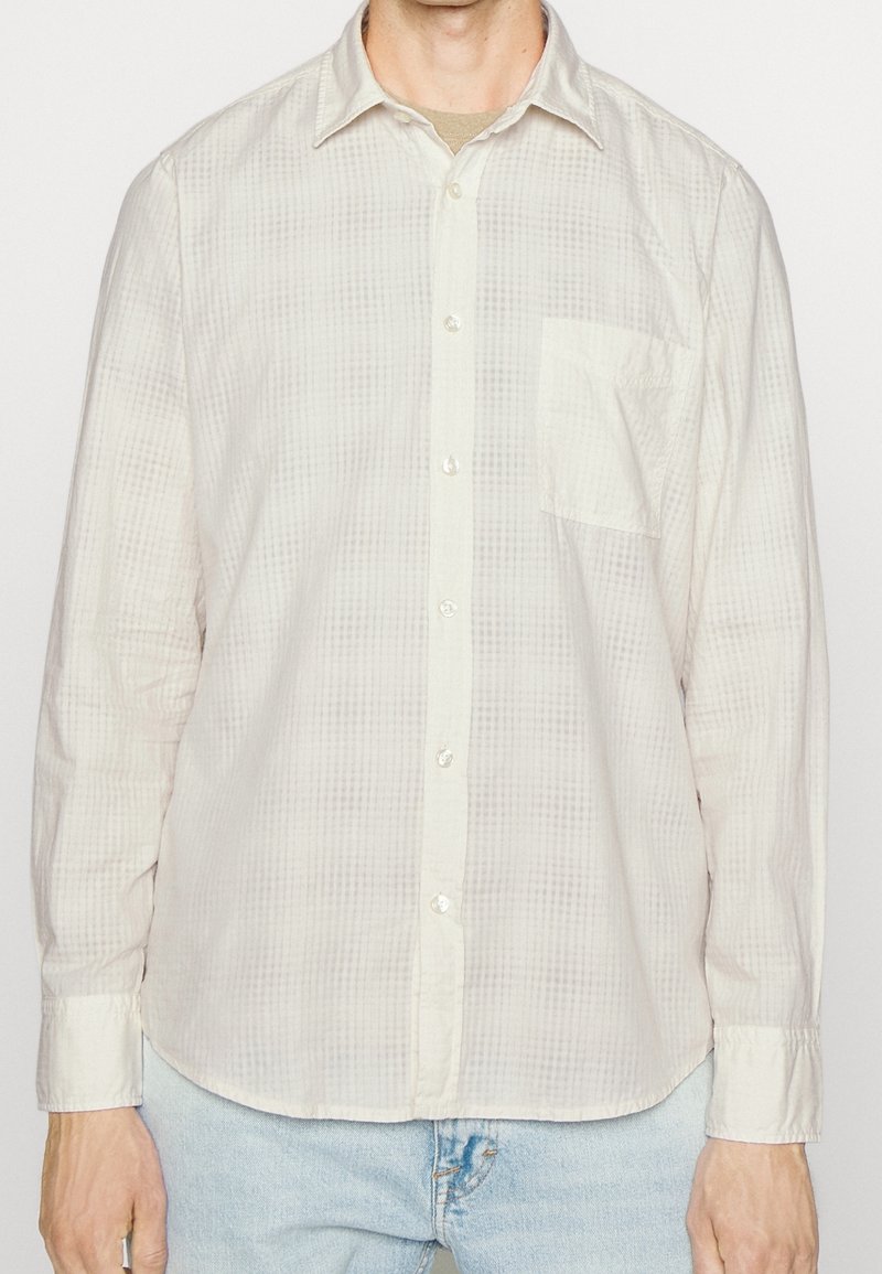 Homme portant une chemise beige clair à manches longues avec un motif à carreaux subtil et une poche poitrine, associée à un jean bleu clair.
