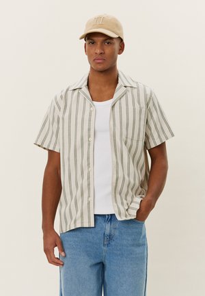 Striped short-sleeve shirt in light beige with green lines, lightweight fabric, buttoned front, single chest pocket, paired with blue jeans.