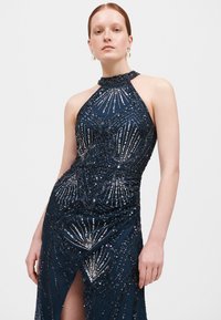 Abito halter neck blu marino con perline, dotato di ricami intricati, corpino aderente e uno spacco alto fino alla coscia, con una finitura lucida e texturizzata.