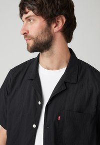 Zwart shirt met korte mouwen, voorzien van een getextureerd patroon, knoopsluiting en één borstzak. Witte T-shirt eronder, stof lijkt lichtgewicht.