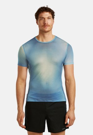 Mężczyzna stojący przodem, ubrany w dopasowaną niebieską koszulkę z krótkim rękawem i czarne spodenki na jednolitym tle.