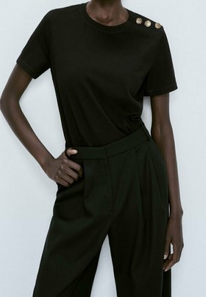 T-shirt noir à manches courtes avec un col rond et trois boutons dorés sur l'épaule gauche, associé à un pantalon noir taille haute.