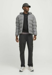 Grå pufferjakke med hætte, sort sweatshirt nedenunder, sorte cargo-bukser og sølvfarvede sneakers. Har lynlåslommer og struktureret stof.