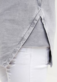Tunic grigia con orlo arrotondato e spacco laterale, realizzata in tessuto morbido. Indossata sopra jeans bianchi strappati, che mostrano texture contrastanti.