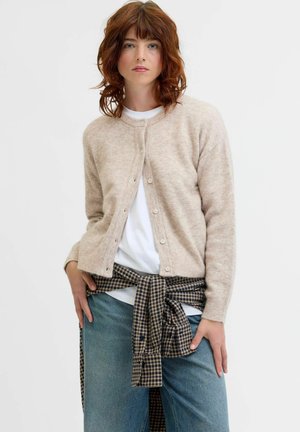 Beige, zachte gebreide cardigan met knoopsluiting, ronde halslijn en ribgebreide manchetten, gedragen over een wit shirt, gecombineerd met blauwe jeans en een geruite blouse.