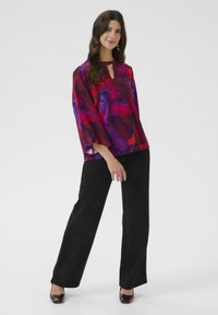 Bluză fluidă, multicoloră în nuanțe de roșu, violet și albastru, cu decolteu tip cheie, asortată cu pantaloni largi negri și tocuri lucioase în nuanță de vișiniu.
