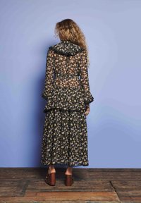 Sieviete ar gariem viļņotiem matiem stāv, vērsta pret zilu sienu, ģērbusies melnā caurspīdīgā ziedu kleitā un brūnās platformas kurpēs uz koka grīdas.