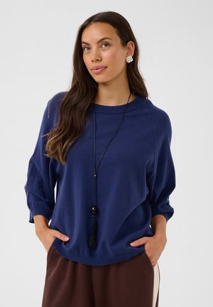 Femme aux longs cheveux bruns porte un pull bleu, un pantalon marron, un long collier pendentif noir et des boucles d'oreilles argentées, debout les mains dans les poches.