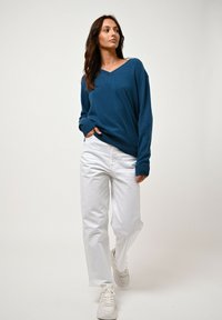 Frau geht mit den Händen in den Taschen, trägt einen blauen V-Ausschnitt-Pullover, weiße Hosen und weiße Sneakers vor einem einfarbigen Hintergrund.