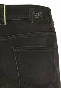 Schwarze Jeanshose mit einer Gesäßtasche und einem Etikett mit "camel active". Der Bund ist mit gelben Nähten gestaltet.