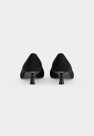 Paire de chaussures noires à bout pointu et talon bas, vues de dos sur un fond clair uni.