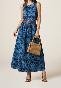 Robe maxi à motifs bleus avec un corsage ajusté et une jupe évasée, ornée d'une ceinture marron. Accessoirisée avec un petit sac en bandoulière tissé.