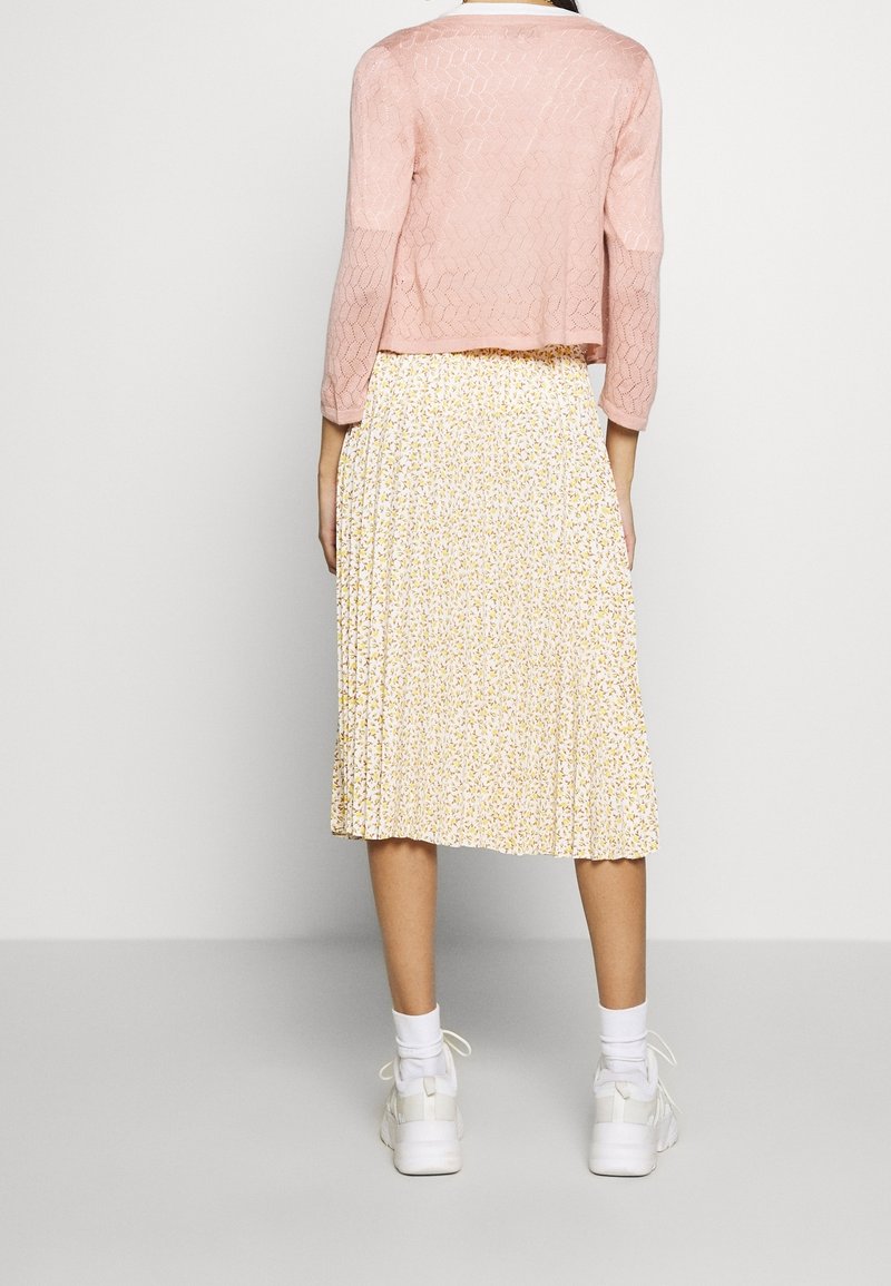 Světle růžový pletený cardigan s tříčtvrtečními rukávy přes bílou plisovanou sukni, která má žlutý květinový vzor, a bílé tenisky.