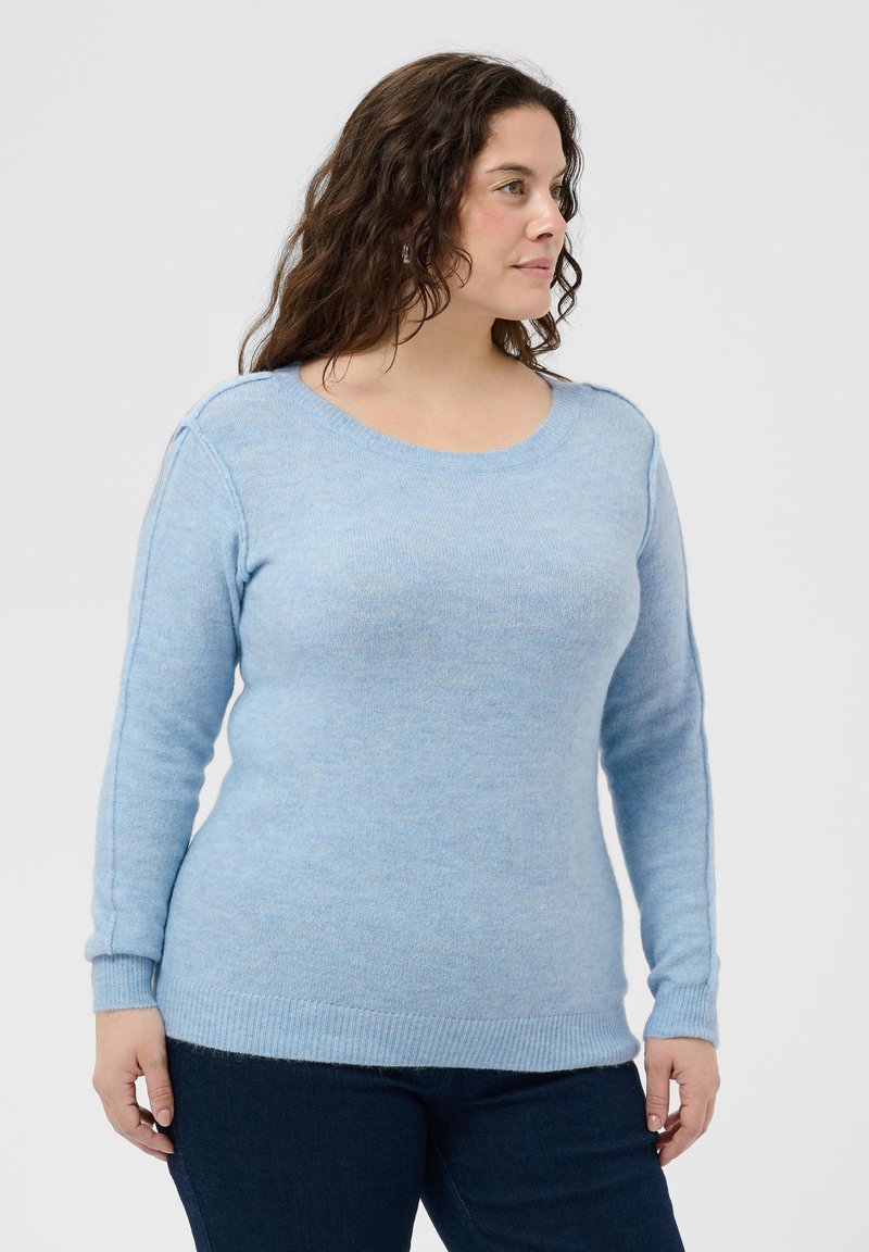 Femme aux cheveux bouclés marron portant un pull bleu clair à manches longues et un pantalon foncé, regardant de côté sur un fond uni.