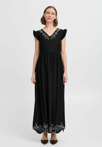 Robe maxi noire avec des manches courtes à volants et un décolleté en V. Présente une broderie de feuilles blanches autour de l'encolure et de l'ourlet. Texture de tissu douce.