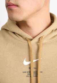 Sweatshirt bege feito de tecido suave, apresentando um capuz com cordões e um logótipo Nike branco. Texto impresso em azul-marinho abaixo do logótipo.