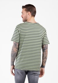 Camiseta verde a rayas con líneas horizontales en azul marino, de manga corta, cuello redondo y un corte relajado. Presenta tatuajes visibles en el brazo del modelo.