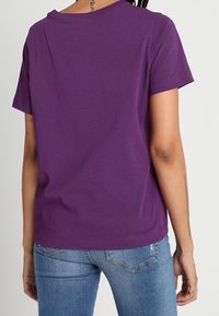 Fialové bavlněné tričko s krátkými rukávy, kulatým výstřihem a rovým lemem. Oblékané s modrými džínami, zobrazený zadní pohled a hladká textura.
