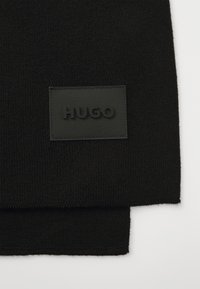 Svart strikket skjerf med en rektangulær teksturert stoffmerke med preget "HUGO"-logo. Enkel design, myk tekstur og ensartet farge.