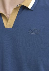 Mornarščina modra polo majica z teksturiranim zaključkom, ki ima kontrastno zlato in belo prožen ovratnik ter vezeno logotip na prsih.