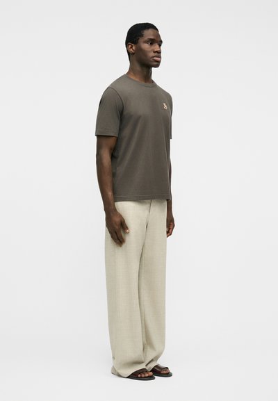 Άνδρας στέκεται στραμμένος προς τα δεξιά, φοράει κοντομάνικο πουκάμισο ελιάς με μικρό έμβλημα, μπεζ παντελόνι φαρδύ στη βάση και σκούρα σανδάλια σε λευκό φόντο.