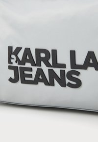Sølvveske med en glatt tekstur og svart preget skrift som leser "KARL LAGERFELD JEANS." Minimalistisk og moderne design.