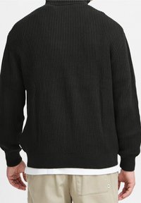 Homme portant un pull en maille côtelée noir sur une chemise blanche et un pantalon beige, vu de dos sur un fond blanc uni.
