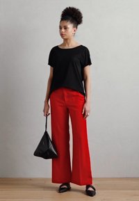 Černé tričko s krátkým rukávem, široké červené kalhoty, černé ploché boty a trojúhelníková černá kabelka. Hladké textury, moderní design.