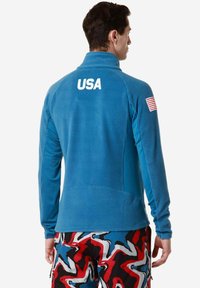 Giacca in pile blu con colletto alto, caratterizzata dalla scritta "USA" in bianco sulla schiena e un patch della bandiera americana sulla manica sinistra.