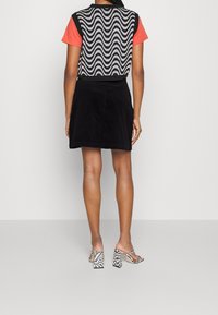 Mujer de pie de espaldas, vistiendo un chaleco de patrón blanco y negro, una camiseta de manga corta coral, una falda negra y sandalias de tacón con patrón.