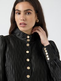 Black pleated blouse with a high collar, featuring gold round buttons along the front and cuffs. Textured fabric adds depth and interest.