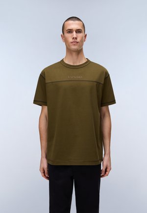 Young man wearing an olive green short-sleeve T-shirt with "NAPAPIJRI" logo across the chest and black pants against a light background.