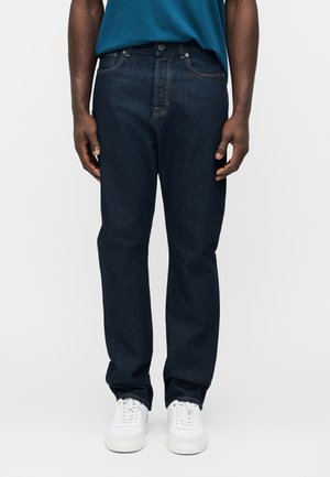 Man wearing dark blue jeans, white sneakers, and a teal shirt, standing against a plain light background.