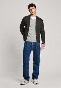 Mörkgrå cardigan för män över en ljusgrå V-ringad tröja, tillsammans med ljusblå jeans och vita sneakers. Enkelt design och modern passform.