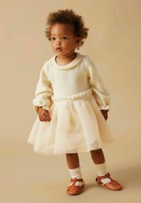 Lys kremfarget genser med ribbestrikket krage, tyllskjørt i blek rosa, og brune sandaler. Strikket tekstur og myke volanger fremhever midjen.