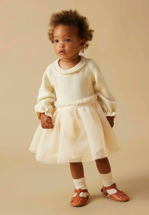 Suéter crema claro con cuello acanalado, falda de tul en rosa pálido y sandalias marrones. La textura de punto y los suaves volantes acentúan la cintura.