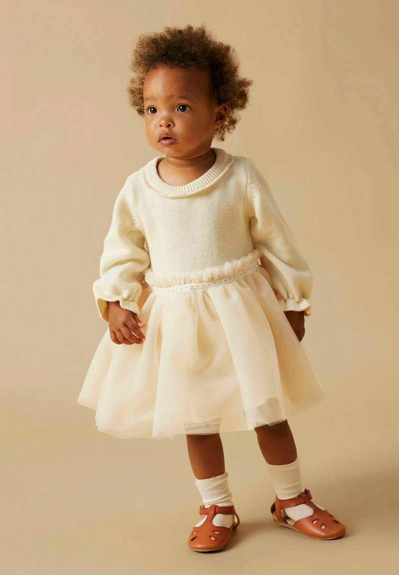 Lys kremfarget genser med ribbestrikket krage, tyllskjørt i blek rosa, og brune sandaler. Strikket tekstur og myke volanger fremhever midjen.