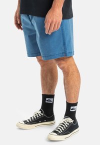 Shorts din bumbac albastru cu buzunare laterale, față plată și margini tivite. Purtați cu adidași negri și șosete cu un logo.