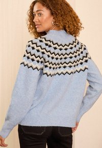 Svetlo modra jopica z geometrijskim vzorcem v črni, belo in sivi barvi na ramah, rebrastimi manšetami in sproščenim krojem.