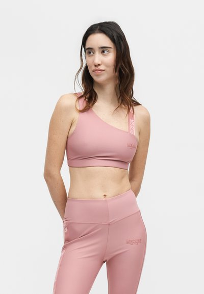 Γυναίκα με μακριά σκούρα μαλλιά φορώντας ένα ροζ αθλητικό σουτιέν με ένα ώμο και αντίστοιχο ψηλόμεσο κολάν, στέκεται με τα χέρια πίσω από την πλάτη.