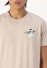 Beige bomull T-skjorte med trykt design av et vinget barn i solbriller på venstre side av brystet. Korte ermer, rund hals.