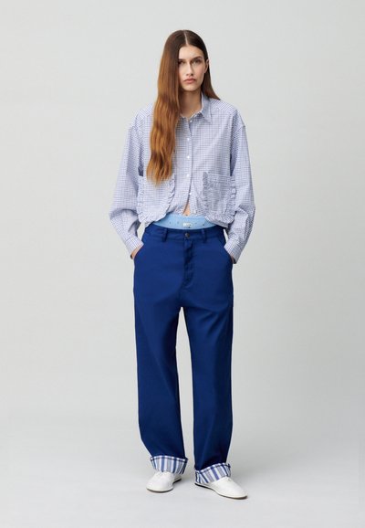 Camicia azzurra a quadri con dettagli arricciati, abbinata a pantaloni blu larghi con polsini a righe e una vita azzurra chiara. Scarpe bianche.