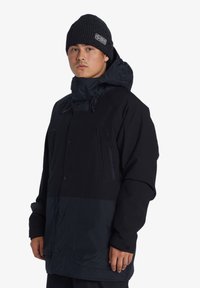Zwarte jas met een donkerblauwe onderkant, voorzien van een capuchon, knoopsluiting en grote zakken aan de voorkant. Draagts met een zwarte gebreide beanie.