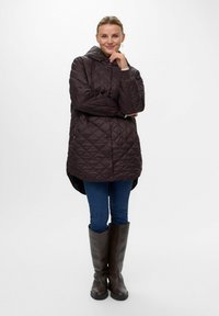 Bruine gewatteerde jas met capuchon, met een ontspannen pasvorm en drukknoopsluiting; gecombineerd met blauwe jeans en bruin kniehoge laarzen.